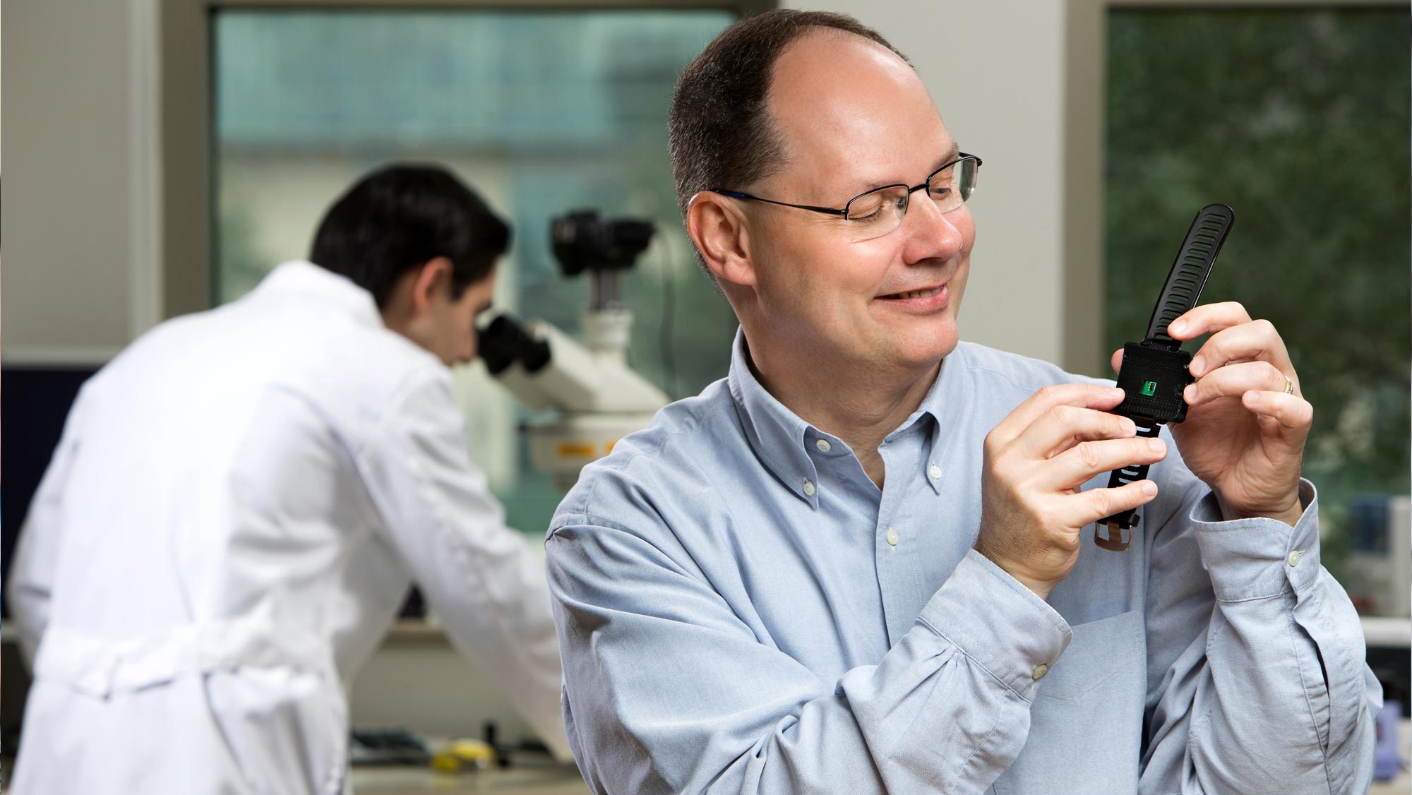 Dr. Gerard Cote smiles as he looks at a wrist device resembling a smartwatch while a researcher who is out of focus works at a microscope behind him.