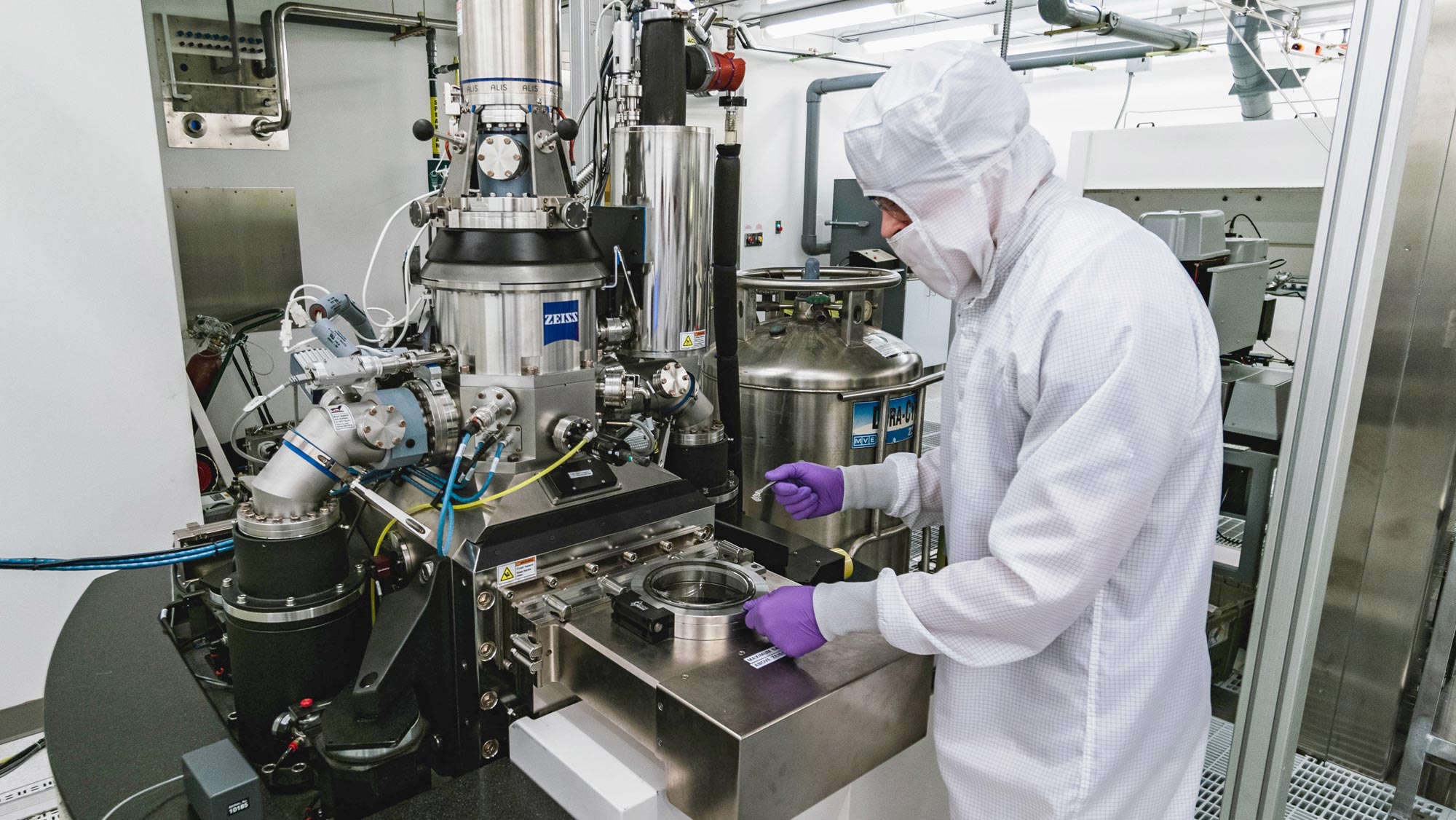 A man or woman covered from head to toe in white safety equipment with only his or her nose and goggles visible, as well as purple latex gloves, holds a small measuring device that resembles a teaspoon in front of silver lab equipment.