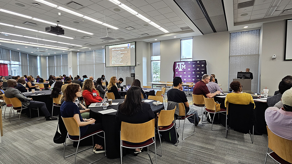A group of people sit around circle tables in the large room looking at a presentation screen. 