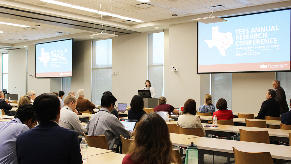 People sit a rows of tables looking at person speaking from a lectern. 