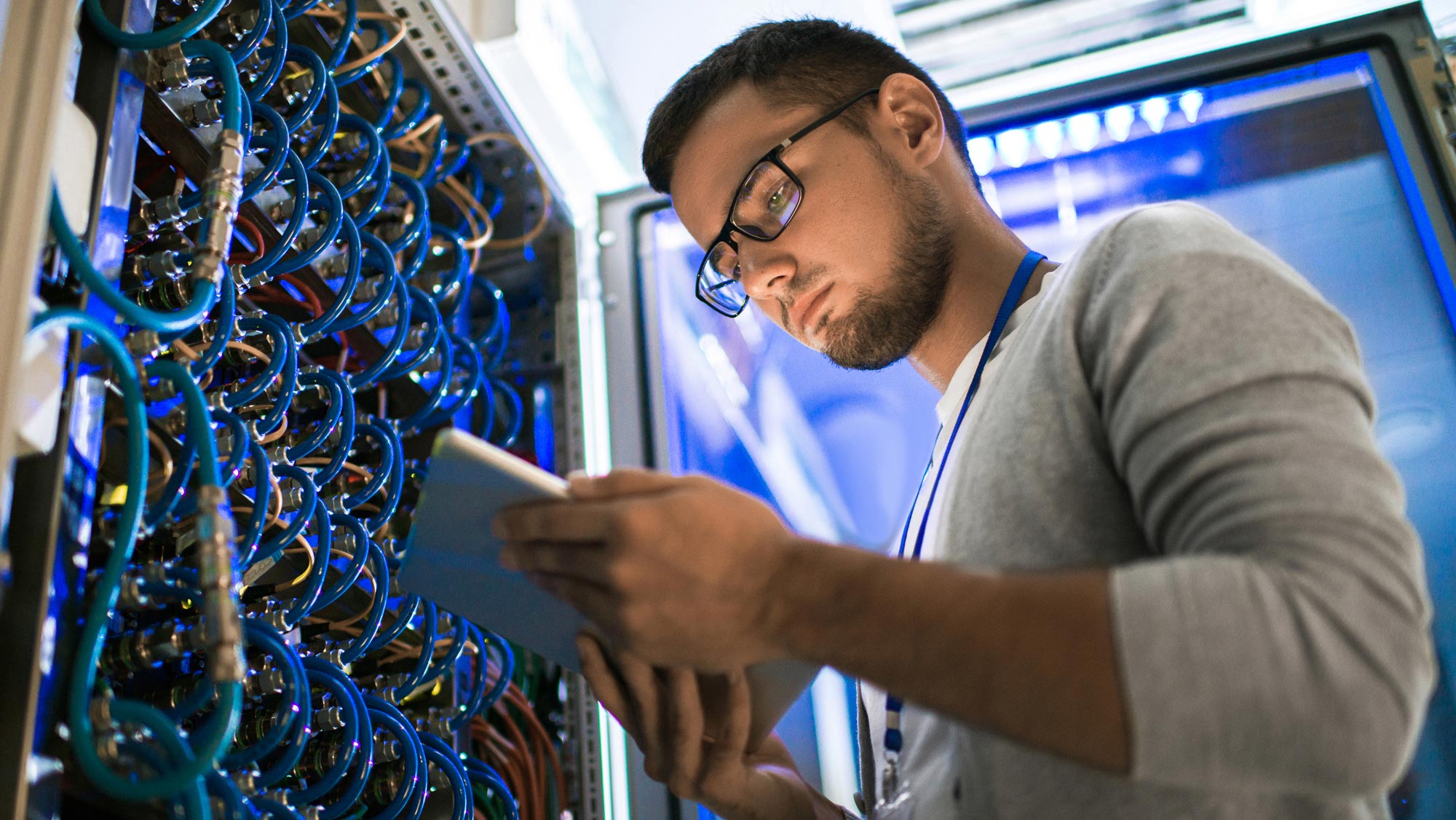Man with dark brown hair and beard, wearing glasses in front of a wall of blue wires as he looks down at a tablet in his hands.