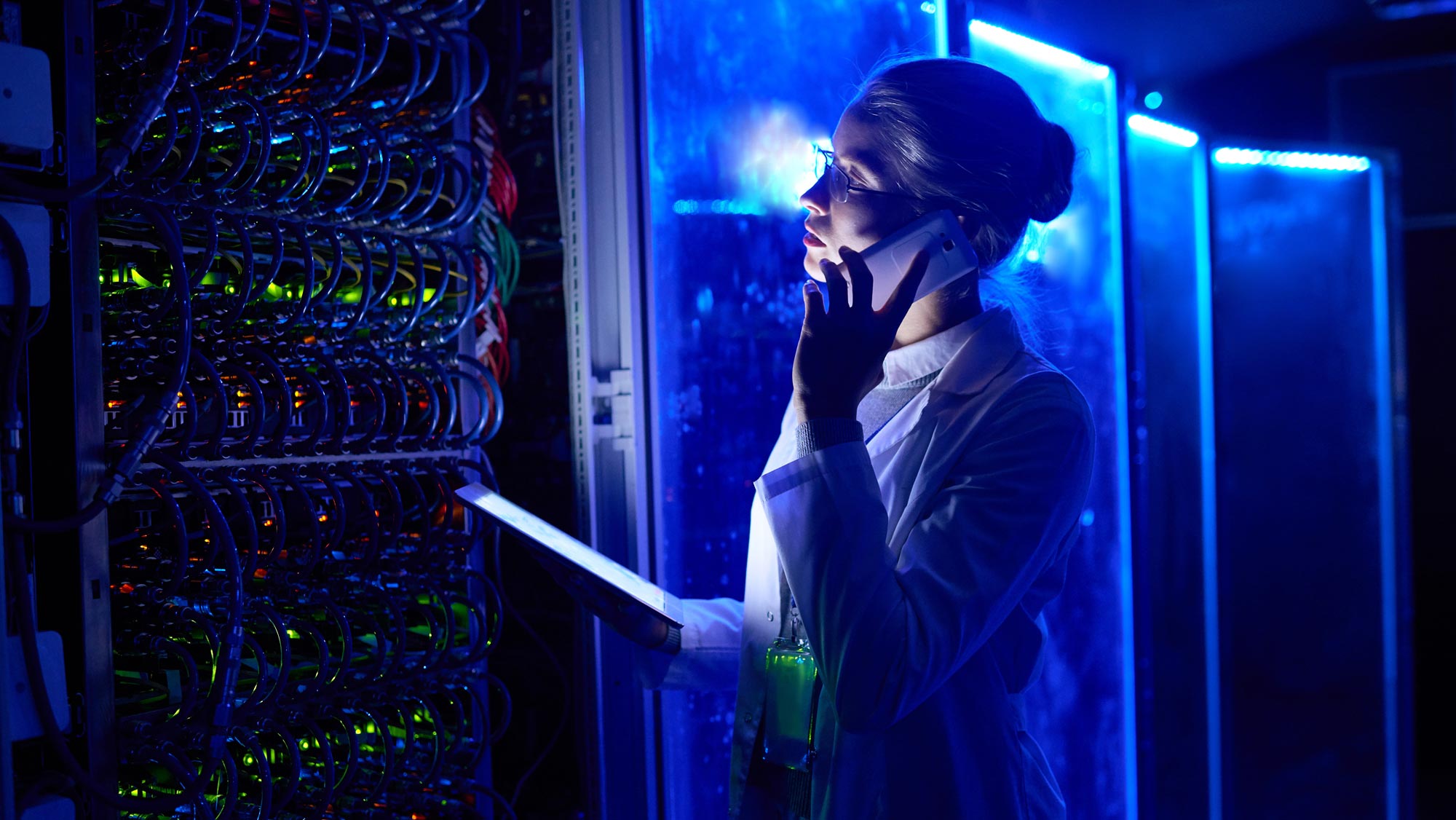 Blonde woman with glasses, hair pulled into a bun, and wearing a white lab coat as she holds a smartphone to her ear and a tablet in her hand while gazing at a panel of blue wires in front of her.