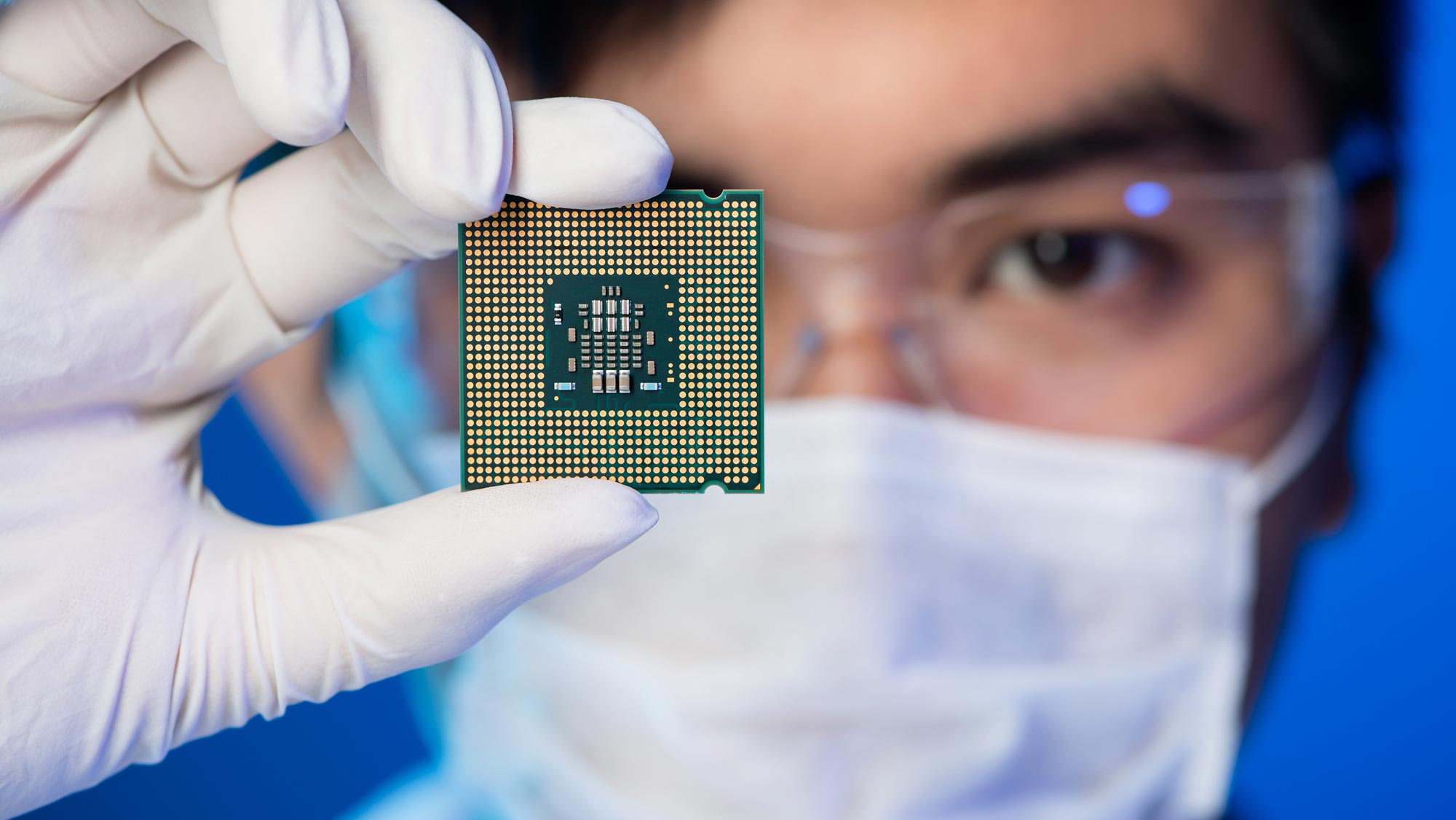 Close-up of a man wearing white gloves, a white face mask, and goggles holding a computer chip.