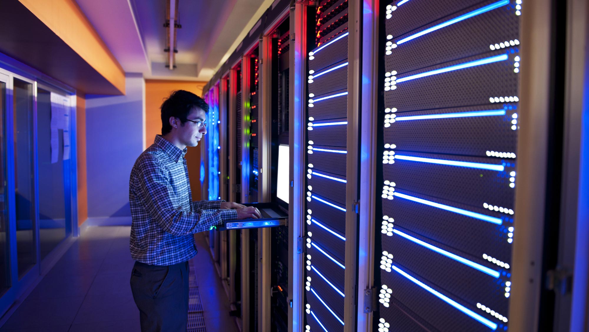 A young man with dark hair and glasses wearing a long-sleeved blue plaid button-down shirt as he works on a laptop in front of computer servers.