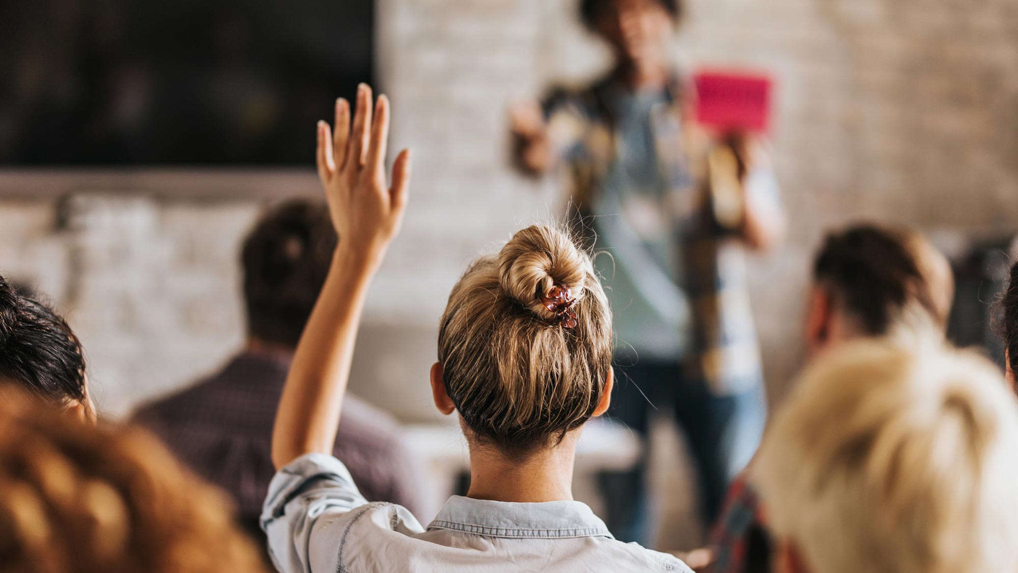 A young woman with her blonde hair pulled into bun faces away from the camera as she raises her hand, waiting for a person at the front of the room who is out of focus to call on her.