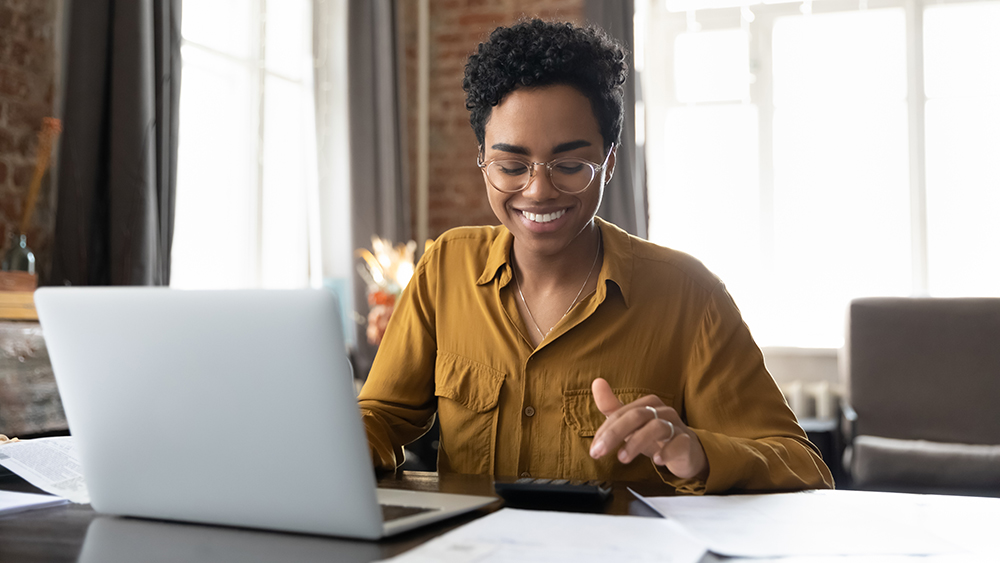 young African American woman wearing glasses and smiling works on a computer laptop while seated at a living room table