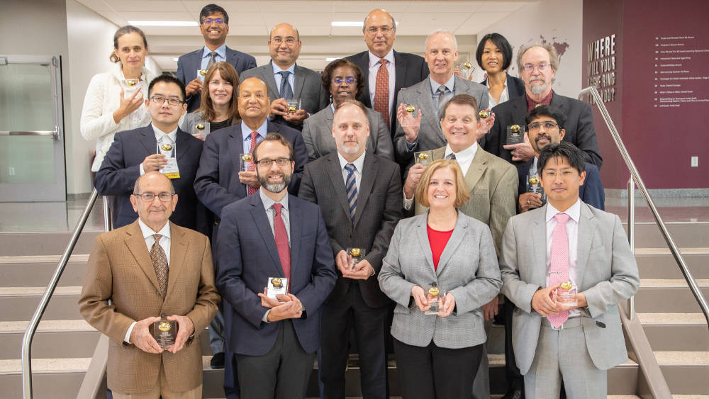 18 people standing on stairs as a group, smiling at the camera while holding "EG Awards," which are awards made of a clear plastic or glass base with a bronze egg mounted on top.