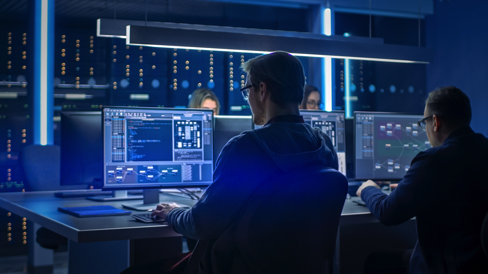 Team of IT programmers working on desktop computers in data center control room. Young professionals writing in sophisticated programming code language.