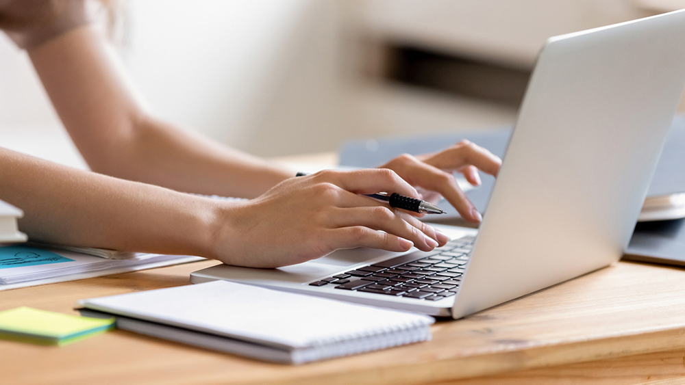 A person's hands and arms holding a mechanical pencil while working on a silver laptop facing away from the camera.