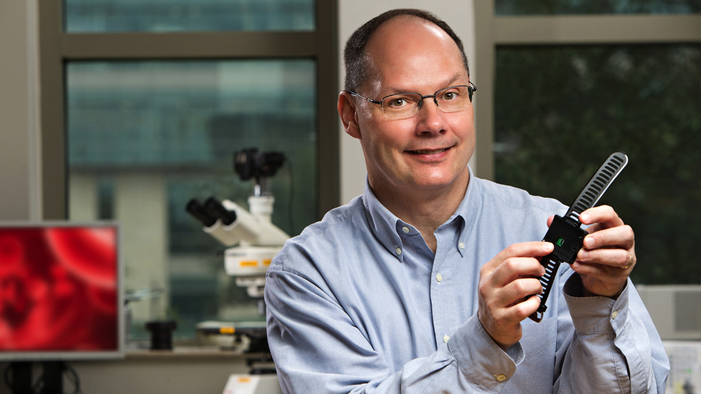Dr. Cote, a smiling man wearing glasses and a button-down blue shirt, points to a smartwatch in his hands. 