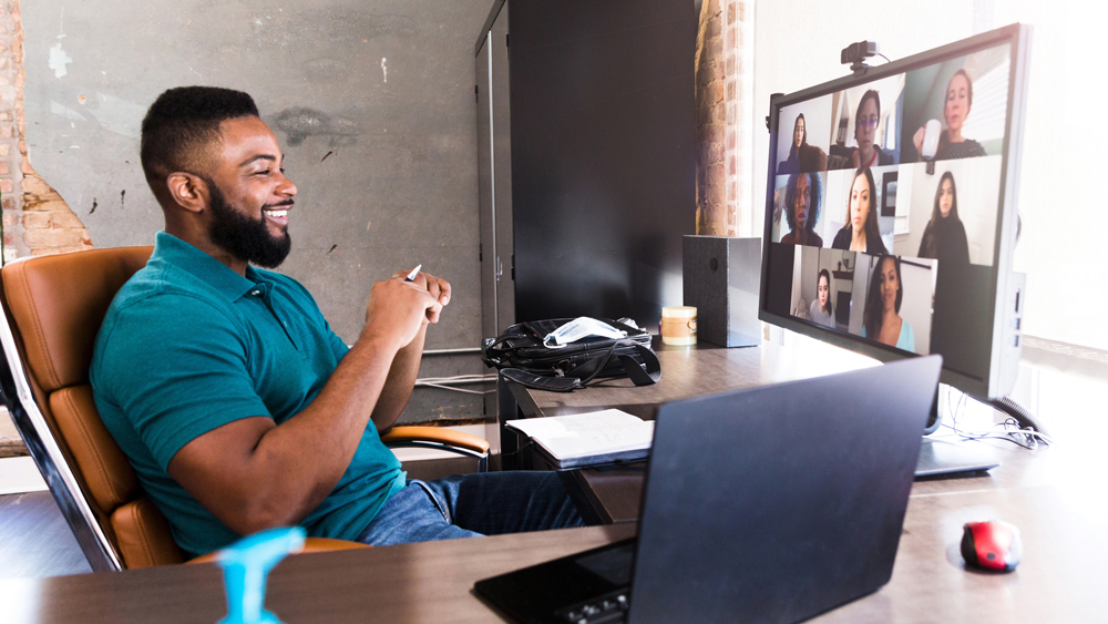 Smiling man with black hair and beard sitting at a desk in front of a computer monitor displaying eight people on a conference call with him.