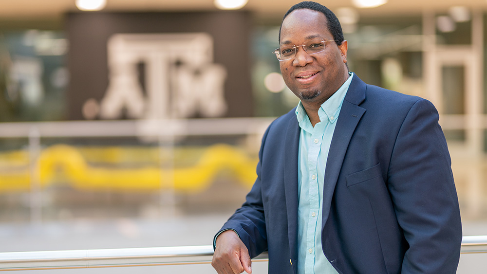Michel Kinsy in a light blue button-down shirt and unbutton navy blazer leans against a railing inside the Zachry Engineering Education Complex.