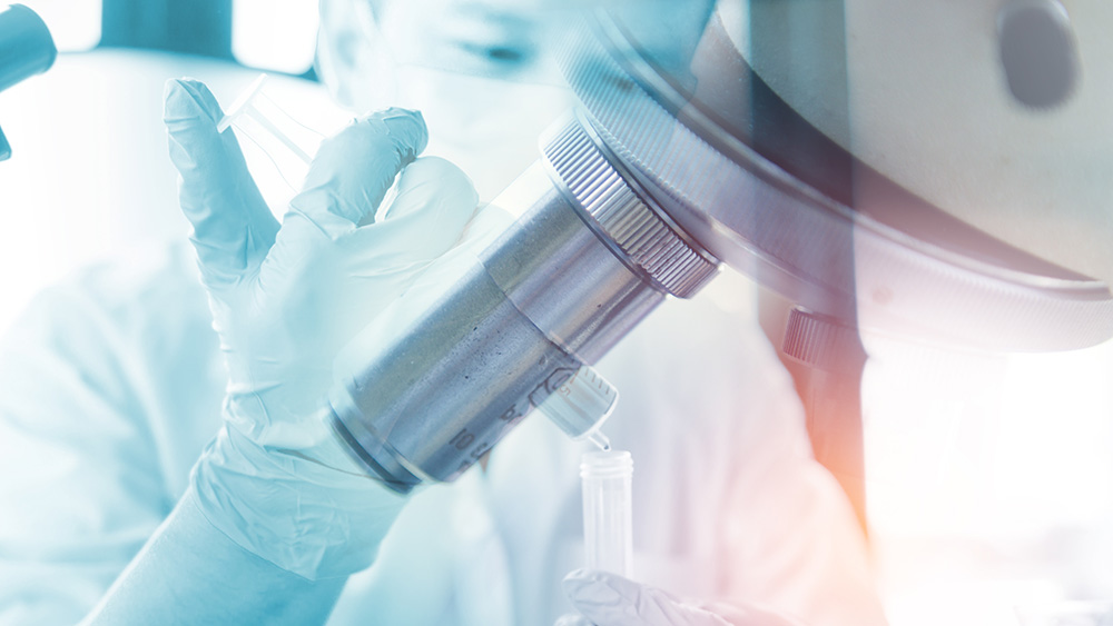 Closeup hand of scientist in a lab holding a syringe squeezing liquid into a tube with a microscope in front.