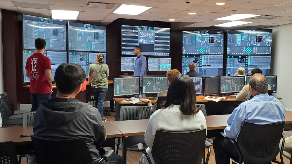 Nine people seated at tables or standing as they observe data on a wall of large computer monitors.