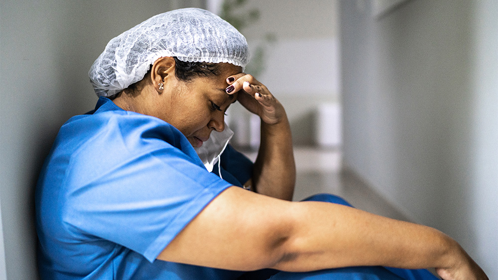 A Black female nurse siting on hallway floor with back against wall, looking down with hand on her head, and a mask partially off her face, in a fatigued state.