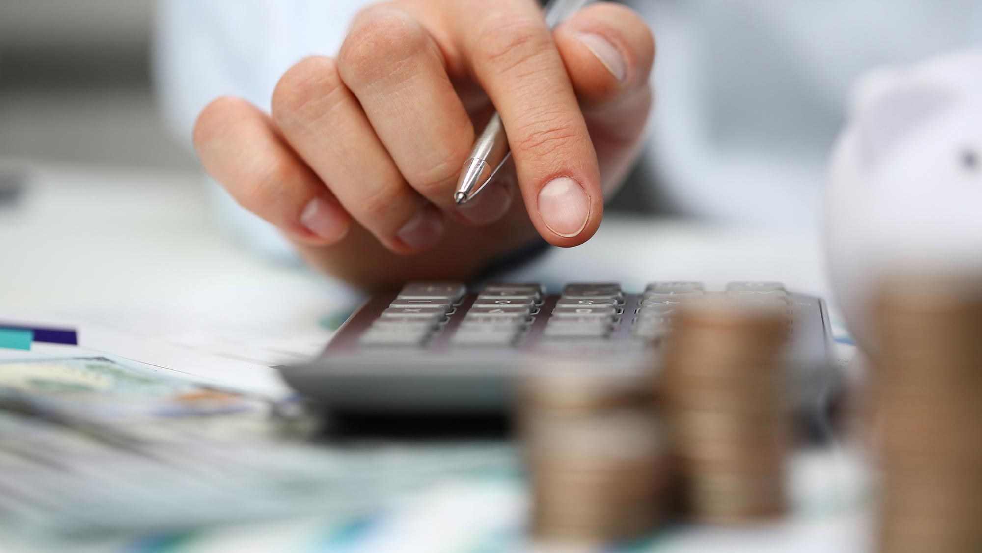 A close-up of a hand holding a silver pen as a well-manicured finger hovers above a calculator.