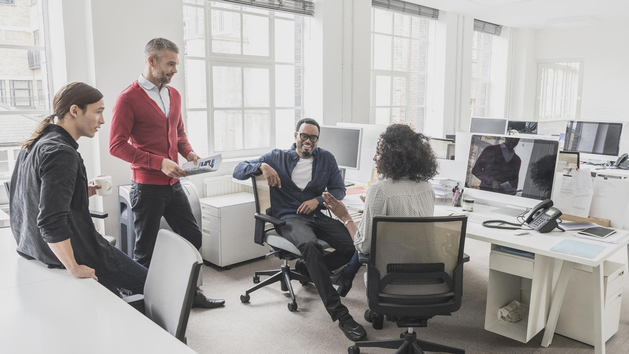 Three men and one woman chat casually in an office full of cubicles, with large windows across one wall.