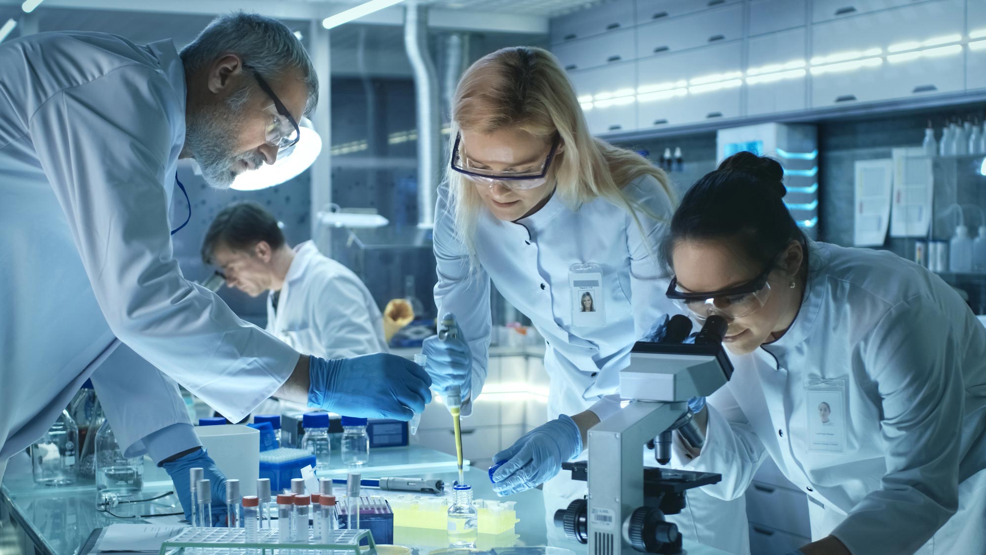 A man with gray hair and a beard, wearing safety goggles, a white lab coat, and blue latex gloves holds a test tube as a woman with blonde hair, wearing safety goggles, blue latex gloves, and a white lab coat fills a pipette with yellow liquid. Next to her, a woman with black hair pulled into a bun, wearing safety goggles and a white lab coat looks through a microscope. Behind them, a man with brown hair and wearing a white lab coat is out of focus as he peers through a microscope. Everyone is in a laboratory setting.