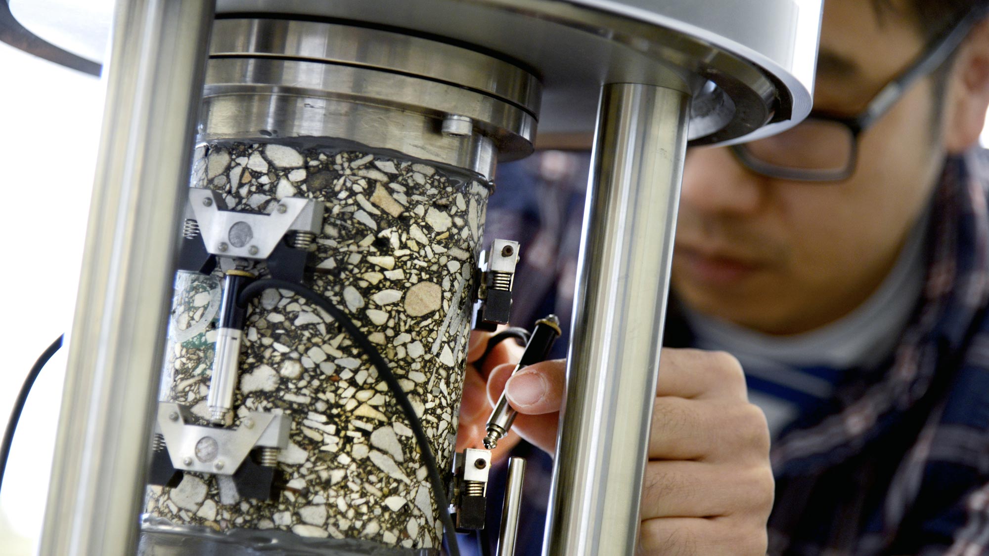 A man with black hair, black glasses, and wearing a blue plaid shirt uses silver lab equipment to work with a cylindrical piece of concrete that appears to contain large fragments of rock in it.