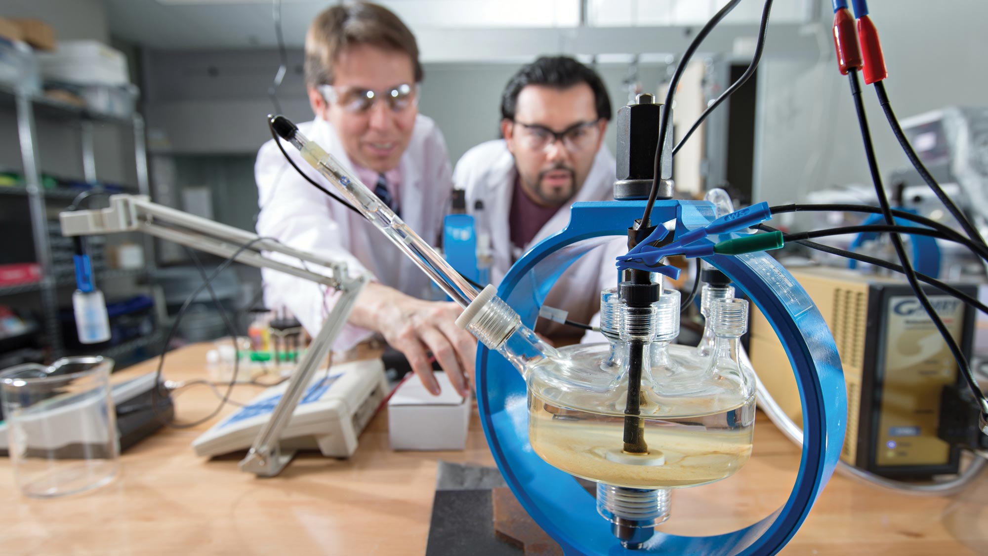 In the foreground is lab equipment that resembles a glass container filled with liquid and surrounded by what appears to be a circle of blue plastic. Out of focus in the background are two male researchers, one with light brown hair and the other with dark hair, both wearing safety goggles and white lab coats.