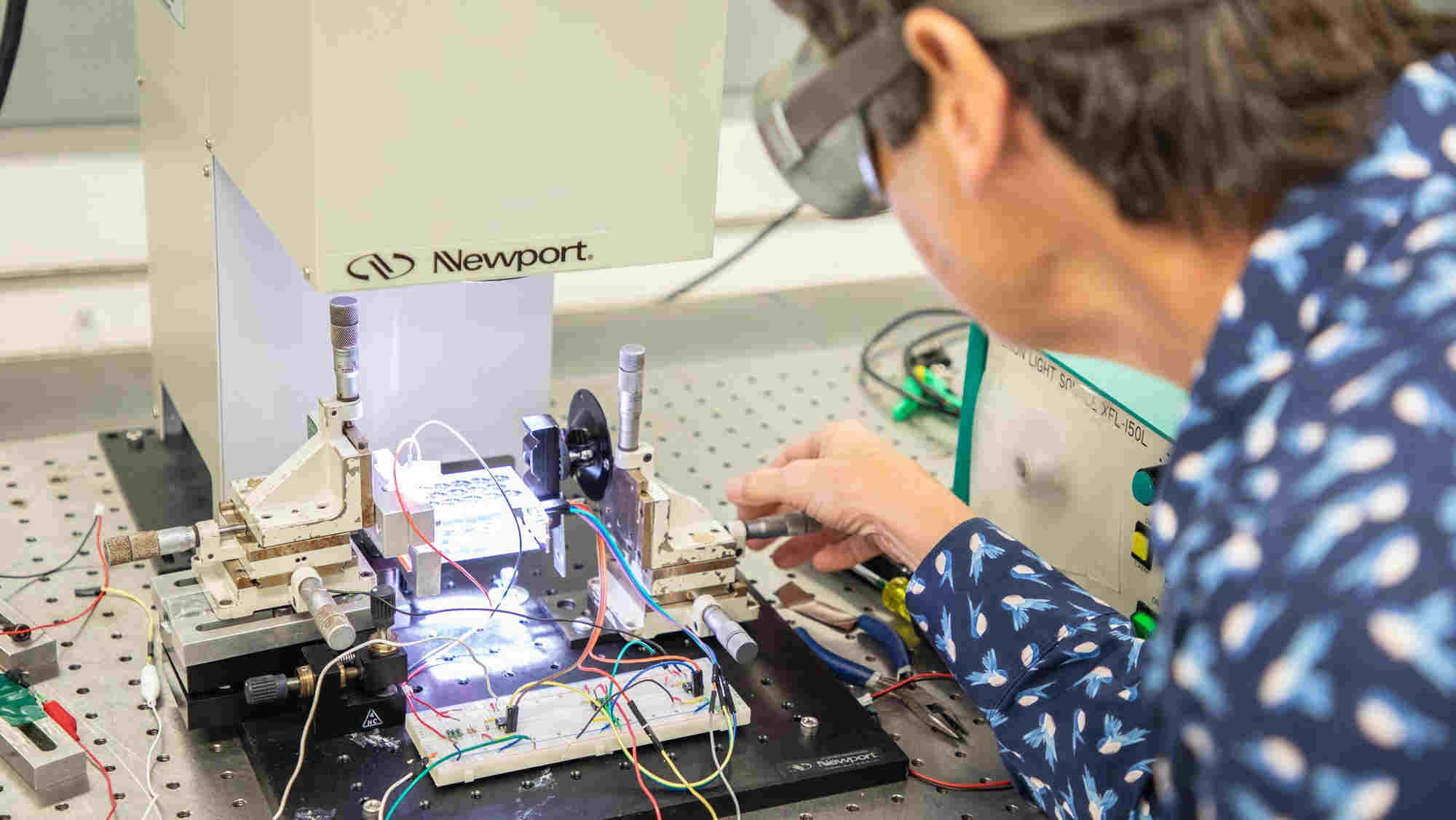 The shoulder and arm of a person wearing safety goggles and a patterned blue shirt with long sleeves reaches for Newport lab machine placed under a light from Light Source XFL-150L machine