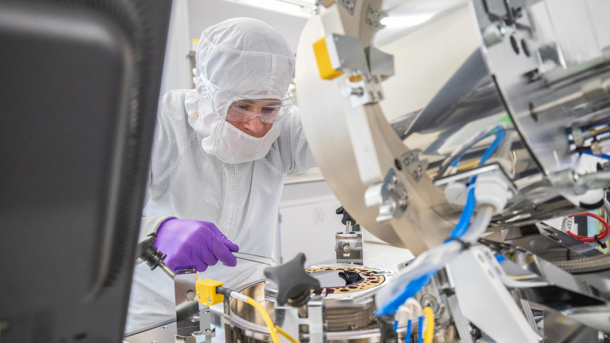 A man or woman covered from head to toe in white safety equipment with only his or her nose, eyes, safety goggles visible, and purple latex gloves visible. He or she holds a large pair of tweezers while working with lab equipment.