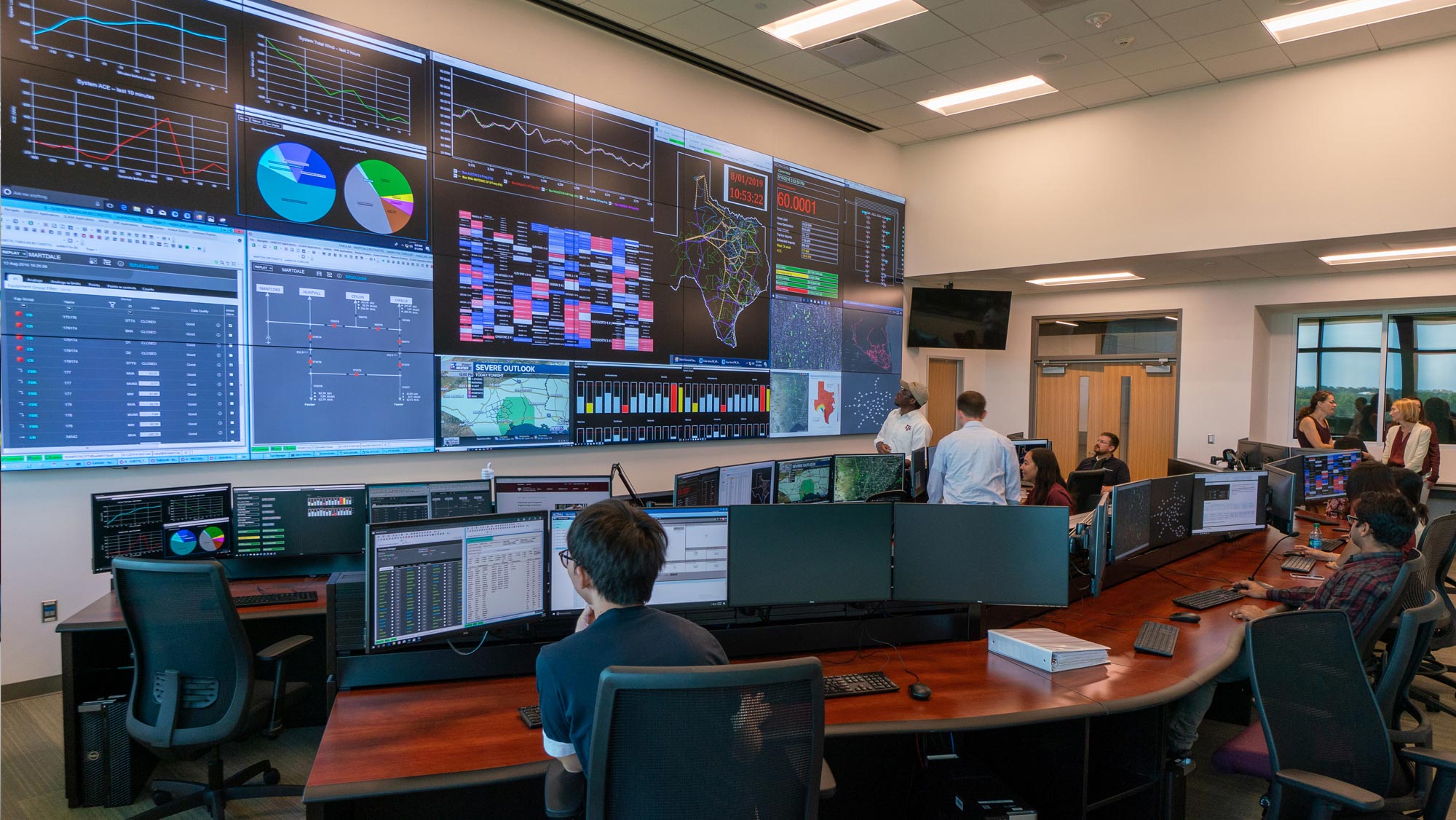 At least eleven people work in a room with two rows of tables that contain computer monitors and keyboards, as well as an entire wall covered with computer monitors, all displaying various charts and graphs.