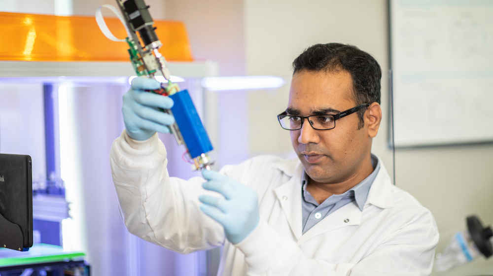 A researcher with black hair and glasses focuses as he works with equipment in a lab setting.