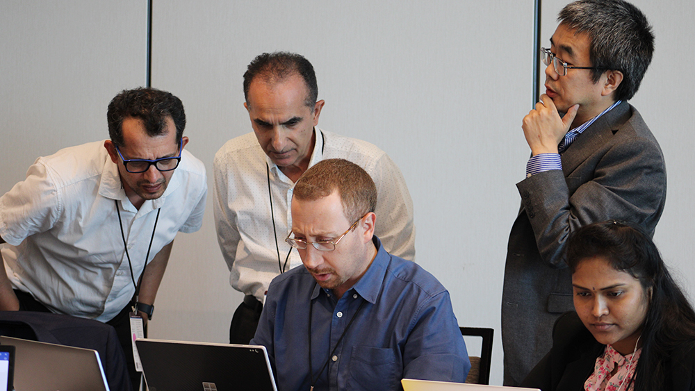 A man and a woman sit at at table working on laptop computers while three men stand behind them looking over their shoulders in discussion. 